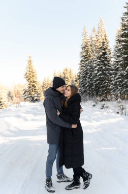 Genç ve aşık bir çift karlı bir yolda sarılıyor. Ormanla çevrili. Kış aile yürüyüşleri.