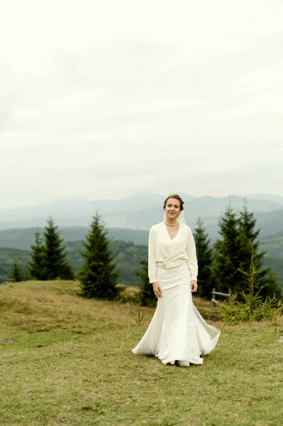 花嫁の肖像画 山や森を背景に 白いウェディングドレスに花と緑の花束を手にした若い女の子が日没時 — ストック写真