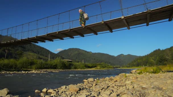 Женщина пересекает подвесной мост через горную реку — стоковое видео