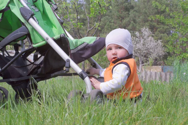 One year old boy sits on the green grass. A little boy is sitting near the stroller. Green baby stroller.