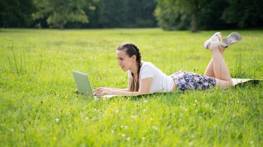 İş dışarıda. Öğrenci kız dizüstü bilgisayarla, bilgisayar tableti de yaz parkında çalışıyor. Çevrimiçi teknoloji ve açık havada bir kadın. Ofis uzaklığı eğitim kavramından kaçmış.