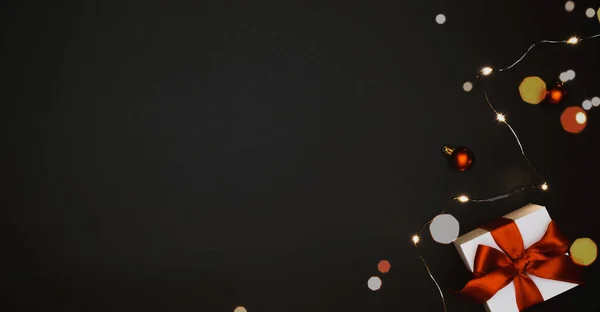 Fondo de invierno de vacaciones. Caja de regalo blanca con cinta roja, bolas de Año Nuevo y guirnalda de luces brillantes en composición navideña en negro para tarjeta de felicitación. Plano, vista superior, espacio para copiar. — Foto de Stock