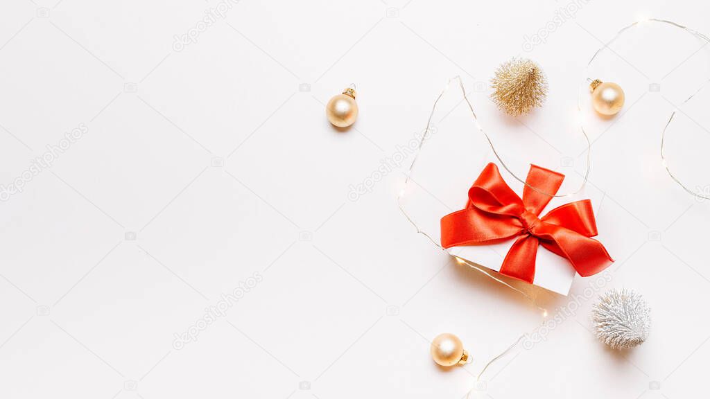 Christmas gift. White gift with red bow, gold balls and sparkling lights garland in xmas decoration on white background for greeting card. Xmas backdrop with space for text