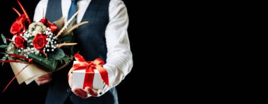 Hediye kutusu eli. Elinde sürpriz hediye kutusu tutan mutlu genç bir iş adamı. Siyah arka planda izole edilmiş bir gül buketi var. Sevgililer Günü, Noel, Yeni Yıl afişi
