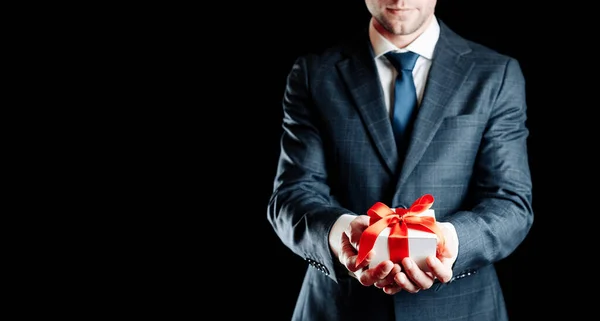Gift man. Happy young businessman holding surprise gift box present with red ribbon isolated on black background. Black Friday, Valentine Day congratulations banner