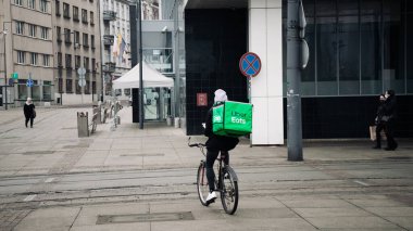 KATOWICE. POLAND - 26 Mart 2021: hızlı restoran teslimatı için bisikletli ya da motosikletli adam. Paket servisi için yemek paketi. Uber yemek dağıtırken mobiletle kurye yiyor.