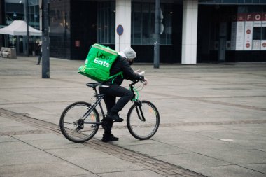KATOWICE. POLAND - 26 Mart 2021: hızlı restoran teslimatı için bisikletli ya da motosikletli adam. Paket servisi için yemek paketi. Uber yemek dağıtırken mobiletle kurye yiyor.