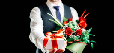 Hediye kutusu eli. Elinde sürpriz hediye kutusu tutan mutlu genç bir iş adamı. Siyah arka planda izole edilmiş bir gül buketi var. Sevgililer Günü, Noel, Yeni Yıl afişi