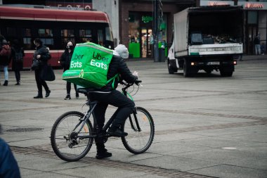 KATOWICE. POLAND - 26 Mart 2021: hızlı restoran teslimatı için bisikletli ya da motosikletli adam. Paket servisi için yemek paketi. Uber yemek dağıtırken mobiletle kurye yiyor.