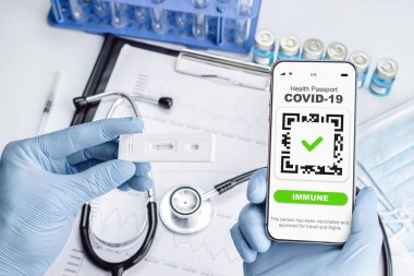 Kapalı pasaport doktoru tutuyor. Doktor steteskop, şırınga ve tıbbi ekipmanlarla birlikte akıllı telefon ekranında dokunulmazlık belgesi.
