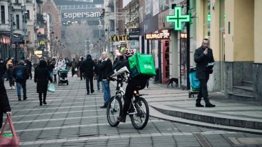 KATOWICE. POLAND - 26 Mart 2021: hızlı restoran teslimatı için bisikletli ya da motosikletli adam. Paket servisi için yemek paketi. Uber yemek dağıtırken mobiletle kurye yiyor.