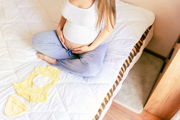 怀孕妇女的婴儿衣服 漂亮的怀孕母亲 穿着黄色的婴儿衣服 孕妇抱着肚子 打包产科医院的包 概念孕产 — 图库照片