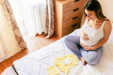 Bebek elbiseli hamile kadın. Sarı bebek elbiseleriyle güzel bir hamile anne. Hamile bir kadın karnına sarılıyor ve hamilelik çantası hazırlıyor. Gebelik, gebelik, doğum