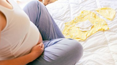 Bebek elbiseli hamile kadın. Sarı bebek elbiseleriyle güzel bir hamile anne. Hamile bir kadın karnına sarılıyor ve hamilelik çantası hazırlıyor. Gebelik, gebelik, doğum