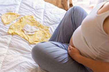 Bebek elbiseli hamile kadın. Sarı bebek elbiseleriyle güzel bir hamile anne. Hamile bir kadın karnına sarılıyor ve hamilelik çantası hazırlıyor. Gebelik, gebelik, doğum