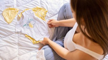 Hamile kadın bebek kıyafetleri. Sarı bebek elbiseleriyle güzel bir hamile anne. Hamile bir kadın karnına sarılıyor ve hamilelik çantası hazırlıyor. Gebelik, gebelik, doğum