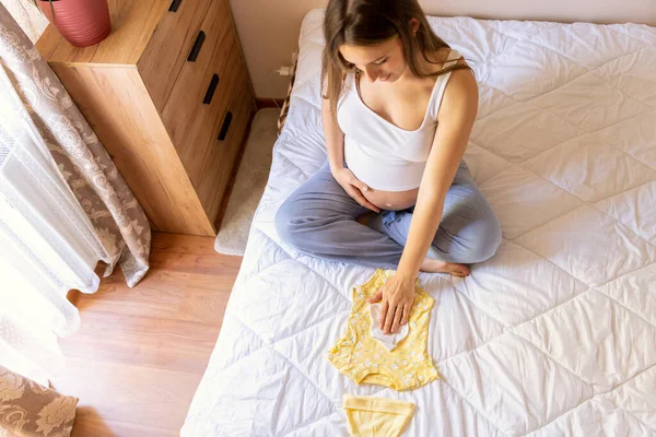 Hamilelik kıyafetleri. Sarı bebek elbiseli güzel hamile bir anne. Hamile bir kadın karnına sarılıyor ve hamilelik çantası hazırlıyor. Gebelik, gebelik, doğum