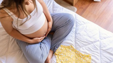 Kadın bebek kıyafetleri. Sarı bebek kıyafetleri tutan mutlu hamile bir anne. Hamile kadın bavul hazırlıyor, doğumevi için çanta. Hamilelik kavramı, annelik, bebek doğumu beklentisi