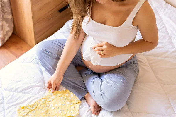 Pregnancy baby clothes. Beautiful pregnant mother with yellow baby clothes. Pregnancy woman hugging belly and packing maternity hospital bag. Concept maternity, pregnancy, childbirth