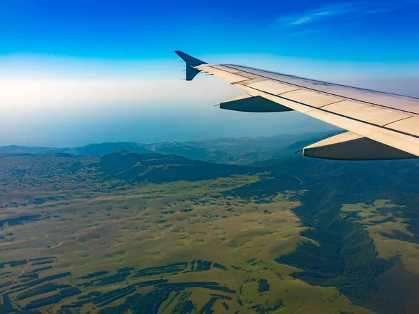 İniş sırasında uçak kanadı, mavi gökyüzü ve yeşil kara manzarası. Uçak penceresi manzaralı. Dünya ve gökyüzü bir uçağın penceresinden görüldüğü gibi.