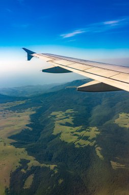 İniş sırasında uçak kanadı, mavi gökyüzü ve yeşil kara manzarası. Uçak penceresi manzaralı. Dünya ve gökyüzü bir uçağın penceresinden görüldüğü gibi.