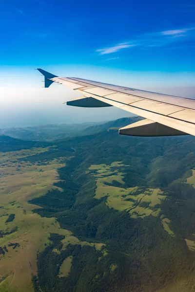 İniş sırasında uçak kanadı, mavi gökyüzü ve yeşil kara manzarası. Uçak penceresi manzaralı. Dünya ve gökyüzü bir uçağın penceresinden görüldüğü gibi.