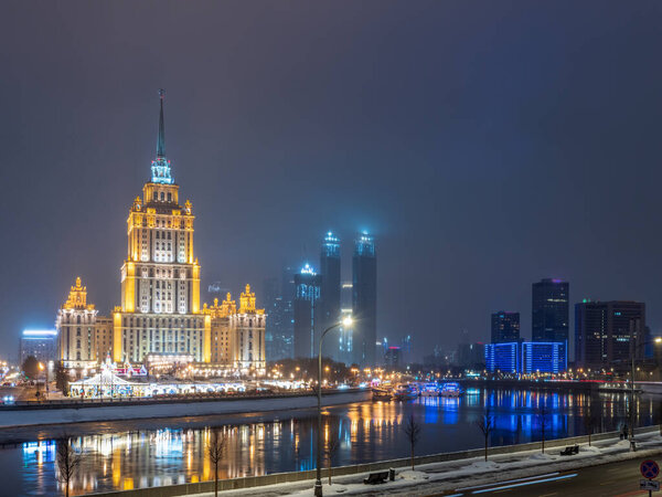 Освещённое высотное сталинистское здание возле реки в зимнюю ночь в Москве, Россия. Зимний городской пейзаж. Перевод надписи на фасаде - Украина.