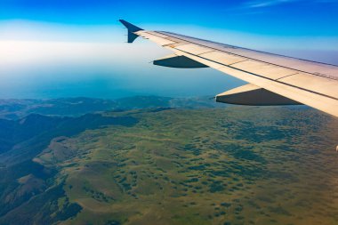 İniş sırasında uçak kanadı, mavi gökyüzü ve yeşil kara manzarası. Uçak penceresi manzaralı. Dünya ve gökyüzü bir uçağın penceresinden görüldüğü gibi.