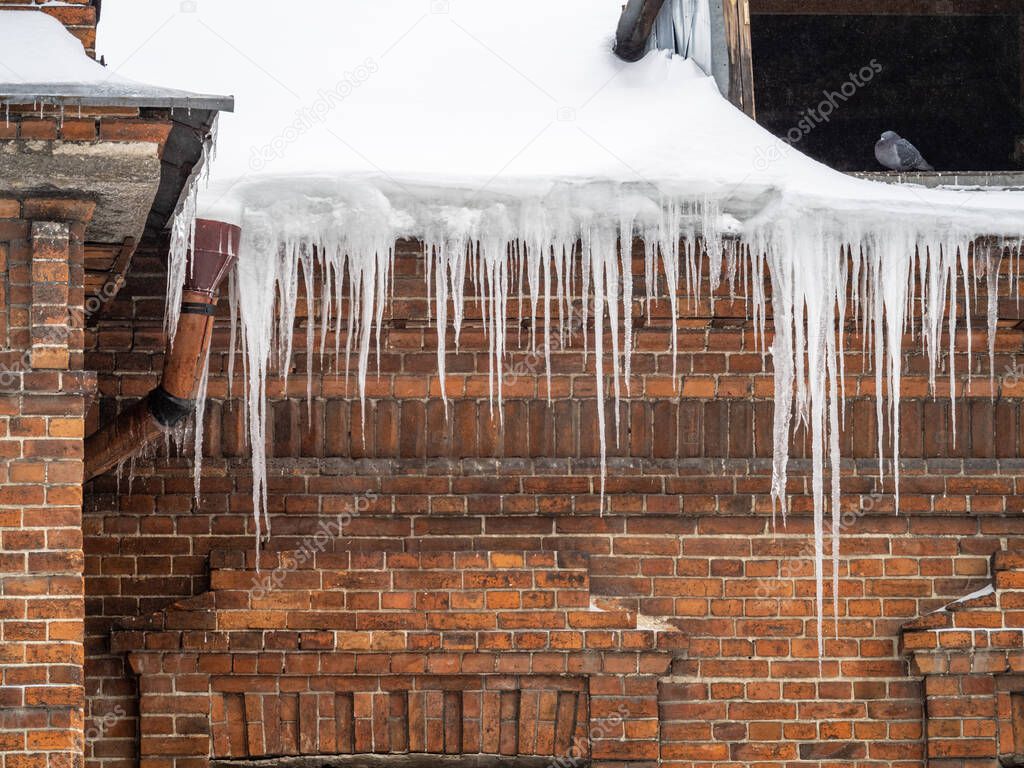 Large icicles hanging from the roof of an old brick building. Roof of old building covered with sharp icicles. Ice stalactite. Poor thermal insulation of the roof leads to the formation of icicles