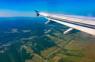 İniş sırasında uçak kanadı, mavi gökyüzü ve yeşil kara manzarası. Uçak penceresi manzaralı. Dünya ve gökyüzü bir uçağın penceresinden görüldüğü gibi.