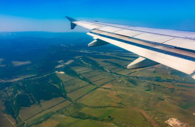 İniş sırasında uçak kanadı, mavi gökyüzü ve yeşil kara manzarası. Uçak penceresi manzaralı. Dünya ve gökyüzü bir uçağın penceresinden görüldüğü gibi.