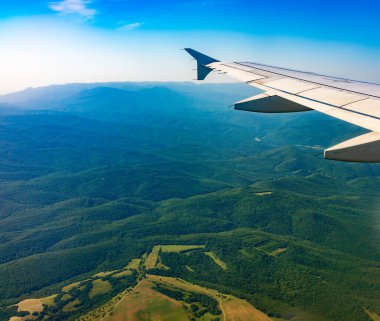 İniş sırasında uçak kanadı, mavi gökyüzü ve yeşil kara manzarası. Uçak penceresi manzaralı. Dünya ve gökyüzü bir uçağın penceresinden görüldüğü gibi.