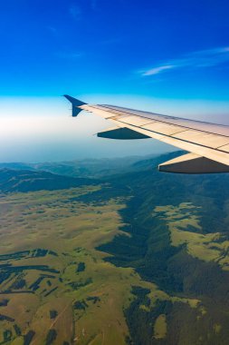 İniş sırasında uçak kanadı, mavi gökyüzü ve yeşil kara manzarası. Uçak penceresi manzaralı. Dünya ve gökyüzü bir uçağın penceresinden görüldüğü gibi.