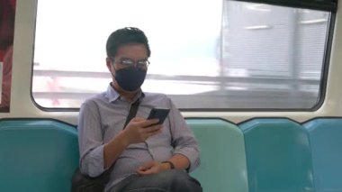 Asyalı işadamı, hareket halindeki bir trenin içinde akıllı telefona bakarken maske takıyor. Maskeli geçiş kavramı.