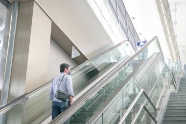 Modern bir binanın içinde yürüyen merdiveni kullanan maskeli bir adam. Seyahat ya da seyahat kavramı.