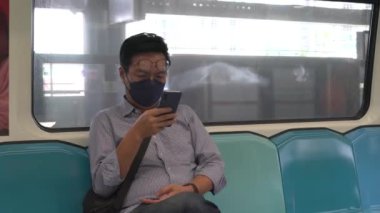 Asyalı işadamı, hareket halindeki bir trenin içinde akıllı telefona bakarken maske takıyor. Maskeli geçiş kavramı.