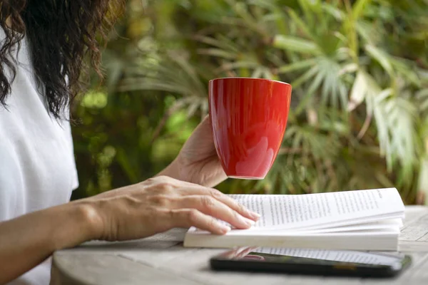 Woman having a cup of coffee and reading a book, close up. Self care or leisure reading concept.