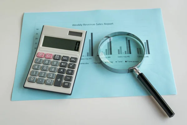 Magnifying glass and calculator on top of sales revenue report with charts. Business performance concept.