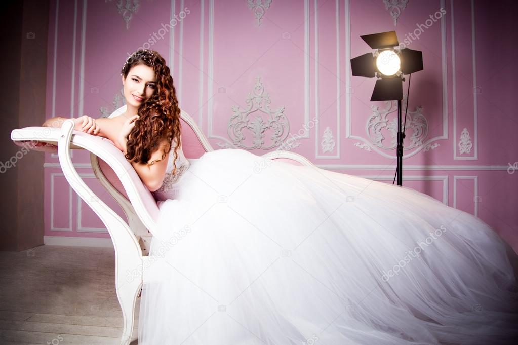 Glamorous photo of a beautiful brunette bride in a luxurious wedding dress lying on pink sofa