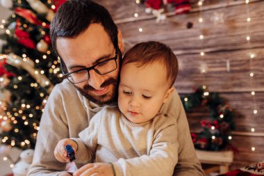 Baba oğluyla Noel ağacı ve hediye kutusuyla dekore edilmiş bir şekilde oturma odasında kucağında oynuyor. Işık rahat bir ortam sağlıyor. Yeni yıl ve mutlu noeller baba ve bebek konsepti