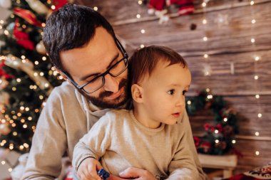 Babası oğluyla Noel ağacı ve hediye kutusuyla dekore edilmiş bir şekilde oturma odasında kucağında oynuyor. Işık rahat bir ortam sağlıyor. Yeni yıl ve mutlu noeller baba ve bebek konsepti