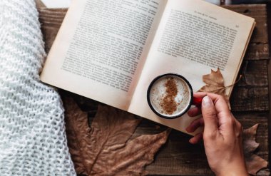 hand of a woman holding cup of coffee while reading a book in autumn