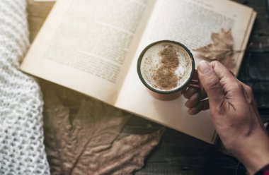 hand of a woman holding cup of coffee while reading a book in autumn