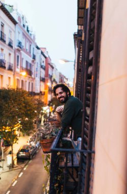 Sıradan bir genç adam, tipik bir Madrid binasının önünde, balkonda gülümsüyor.