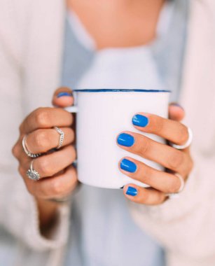 Mavi tırnaklı, yüzüklü, elinde kahve ya da çayla bembeyaz bir kupa tutan genç bir kadının ellerine yakından bakın.