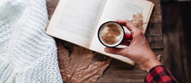 Sonbaharda kitap okurken elinde bir fincan kahve tutan kırmızı ekose ceketli bir kadının üst görüntüsü.