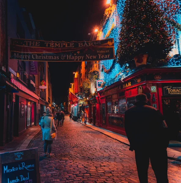 DUBLIN, IRLAND, DECEMBER 24, 2018: Människor promenader i Temple Bar historiska distrikt, ett kulturellt kvarter med livligt nattliv. Nightscene av baren, full av ljus och dess typiskt irish pub utseende. — Stockfoto