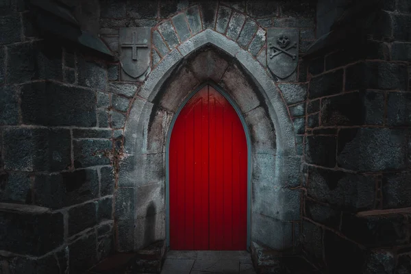 Puerta de madera roja puntiaguda asustadiza en un edificio de pared de piedra vieja y húmeda con cruz, cráneo y huesos a ambos lados. Concepto misterio, muerte y peligro. Imagen De Stock