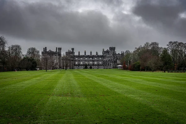 KILKENNY, IRELAND, 23 ARALIK 2018: Kilkenny Şatosu dramatik bulutlu bir günde yeşil çimenlerin üzerinde kuru yapraklarla büyük bir bahçeden görüldü