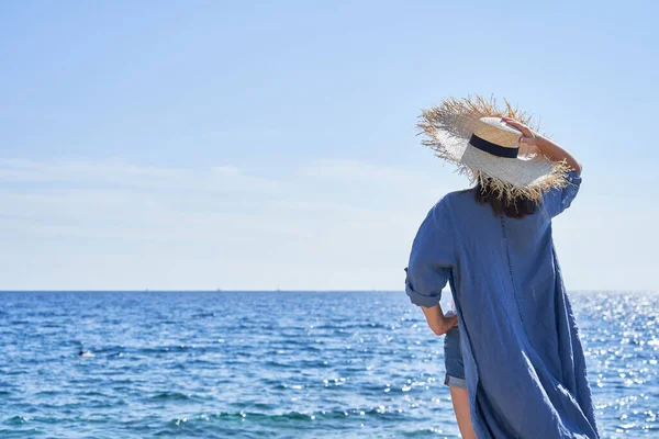 Arkadan bir kadın görülüyor. Rüzgarda çırpınan giysiler. Hasır şapka. Adria Denizi 'nin önündeki kayalıklarda duruyor..